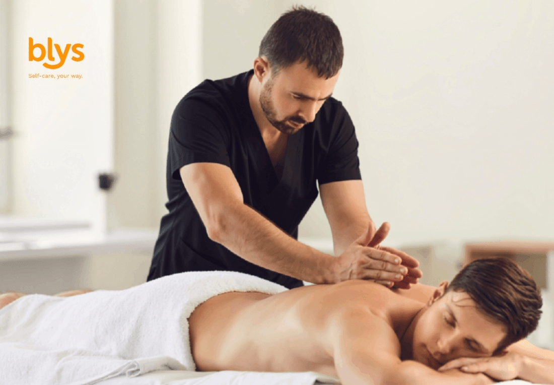 A remedial massage therapist gently presses a client’s back as he lies face down under a white towel in a bright room, conveying calm and care.