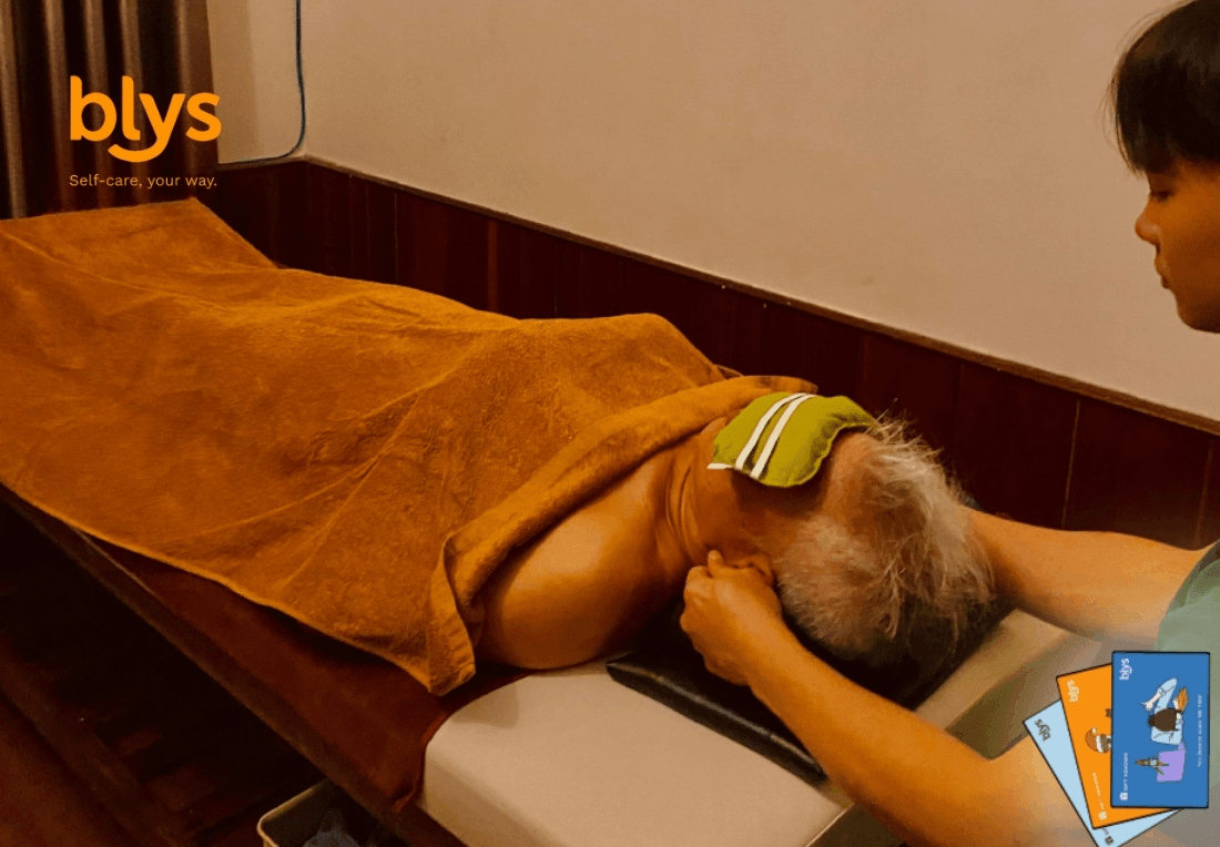 A home care worker massages a reclining client covered by an orange towel, placing a striped cloth on their head; the warm-lit treatment room feels calm and soothing.