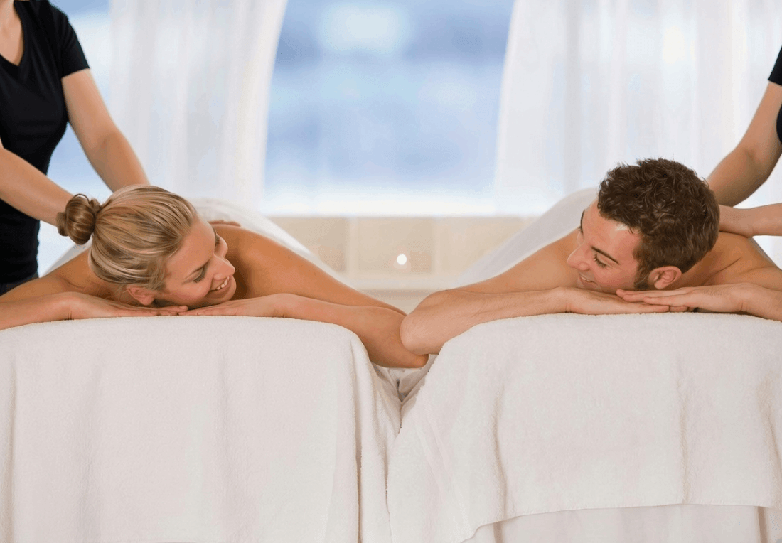 A relaxed couple enjoys a side-by-side mobile massage on white-covered tables, smiling at each other as therapists work in a bright, serene room.