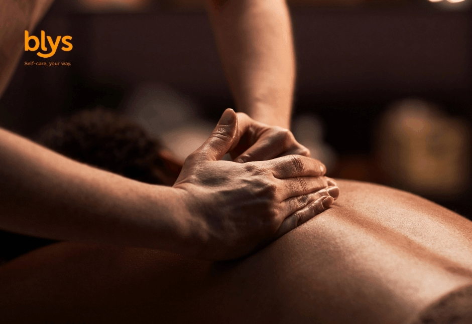 A remedial massage therapist presses both hands into a client’s back during a warm, dimly lit massage; the close-up conveys calm, care, and relaxation.