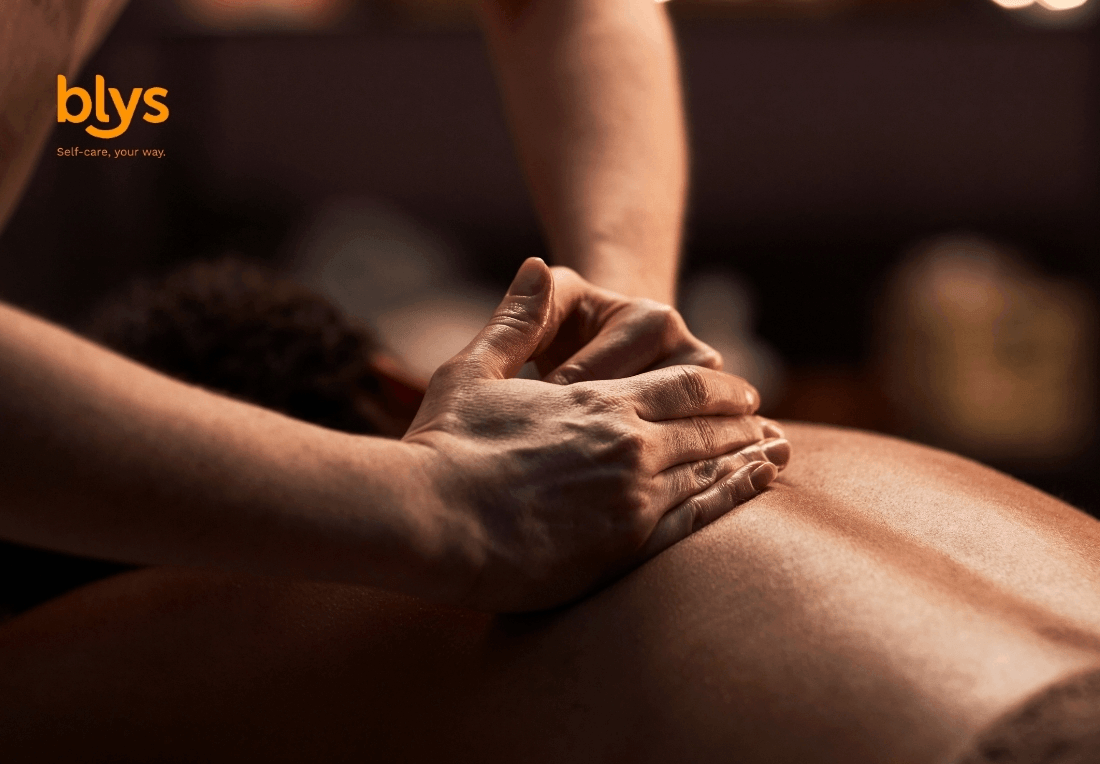 A remedial massage therapist presses both hands into a client’s back during a warm, dimly lit massage; the close-up conveys calm, care, and relaxation.
