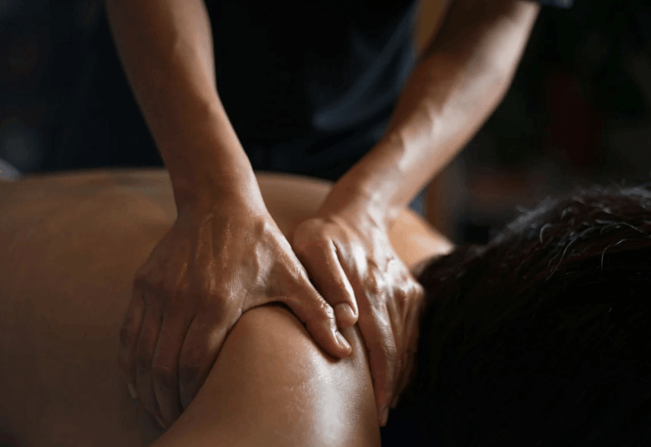 A remedial massage therapist presses both hands into a person’s bare upper back and shoulder in a dim room, conveying relaxation and relief.