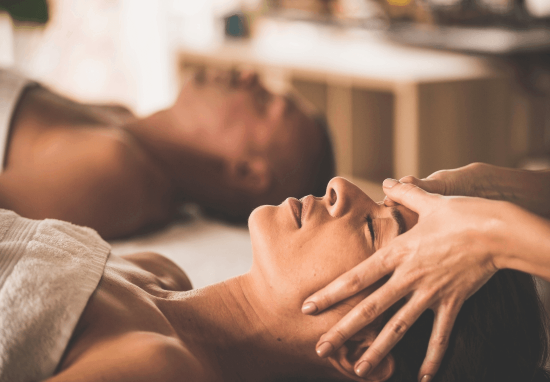 A relaxed couple receives a gentle head and face massage at a room, with a therapist’s hands at a woman's temples and jaw; warm light creates a soothing, peaceful mood.