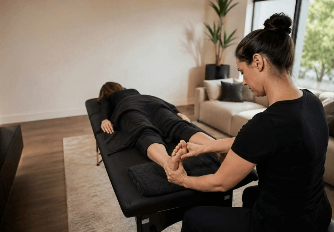 A reflexologist gently presses a client’s foot on a padded treatment table in a calm, sunlit room, creating a relaxed wellness setting.