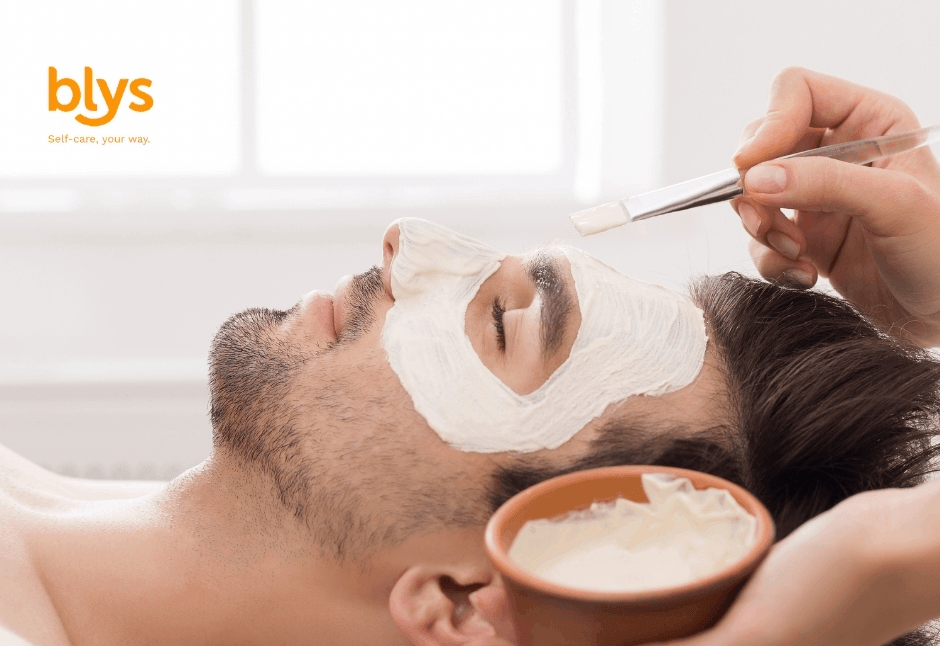 Relaxed man receives a soothing white facial mask at a spa as an esthetician applies cream with a brush; skincare bowl nearby.