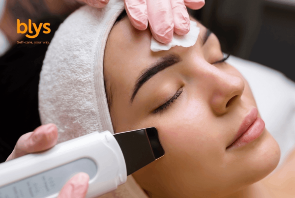 Woman relaxes during a facial cleansing treatment as a gloved esthetician uses an ultrasonic skin spatula on her cheek; soothing spa setting.