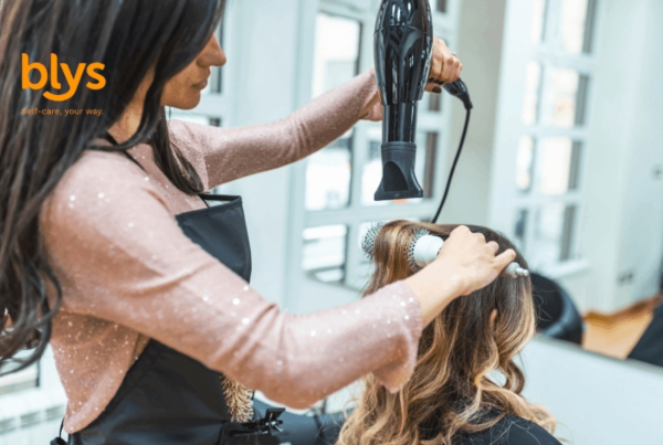 Beauty therapist blow-dries a client’s wavy hair with a round brush in a bright room, creating a polished look in a professional, upbeat setting.