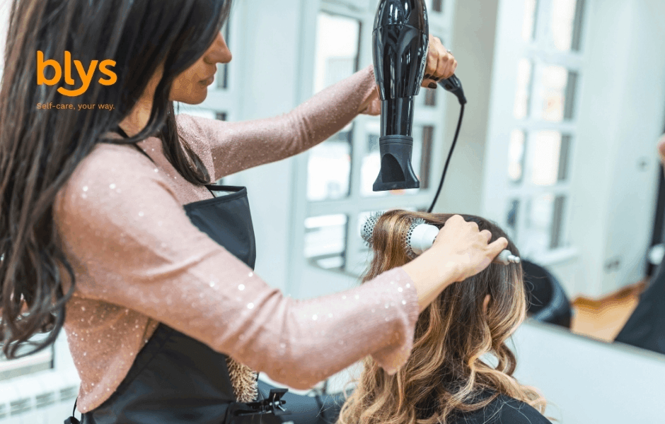 Beauty therapist blow-dries a client’s wavy hair with a round brush in a bright room, creating a polished look in a professional, upbeat setting.
