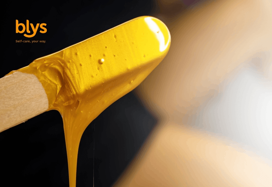 Close-up of a wooden waxing spatula coated in glossy golden hair-removal wax, dripping against a dark background; blys logo and tagline appear above.