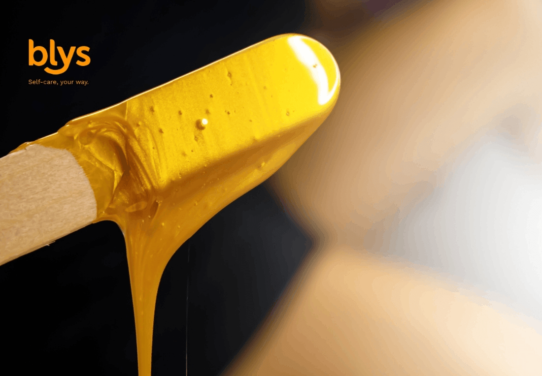 Close-up of a wooden waxing spatula coated in glossy golden hair-removal wax, dripping against a dark background; blys logo and tagline appear above.