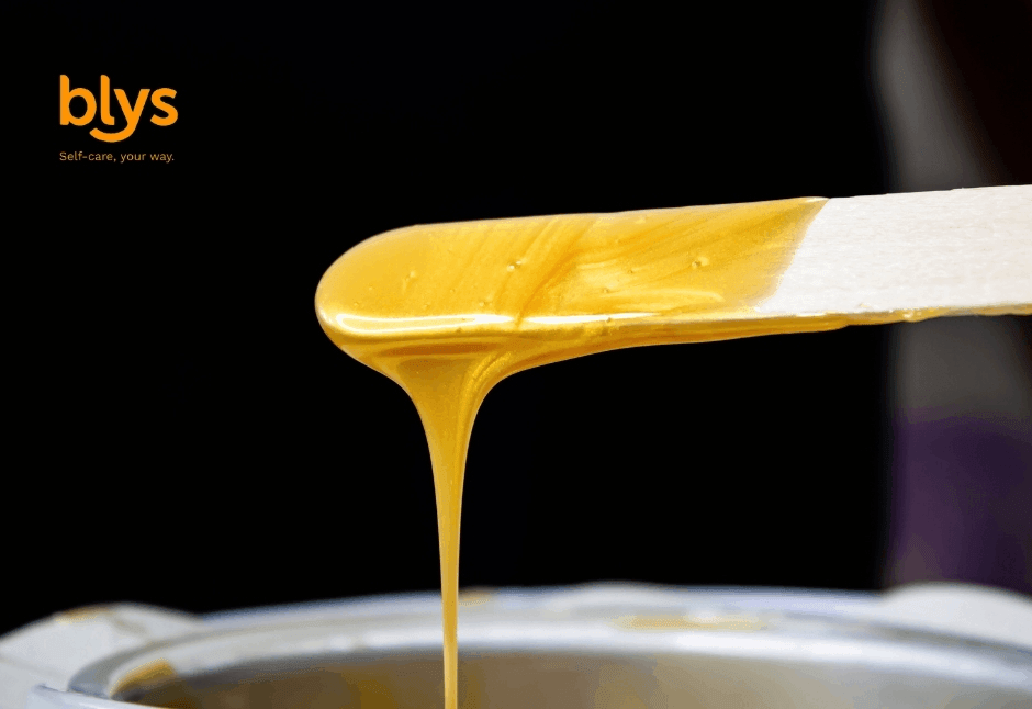 Close-up of golden skincare wax dripping from a spatula into a metal bowl against a black background; warm, glossy, professional tone.