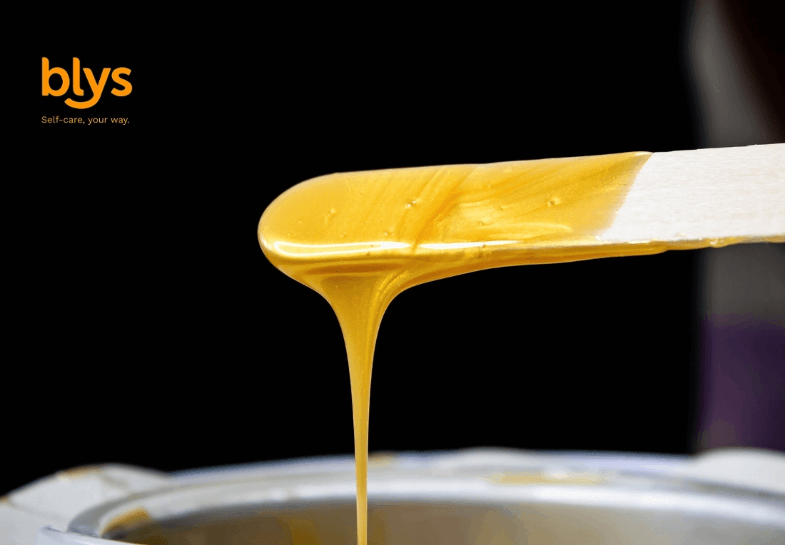 Close-up of golden skincare wax dripping from a spatula into a metal bowl against a black background; warm, glossy, professional tone.