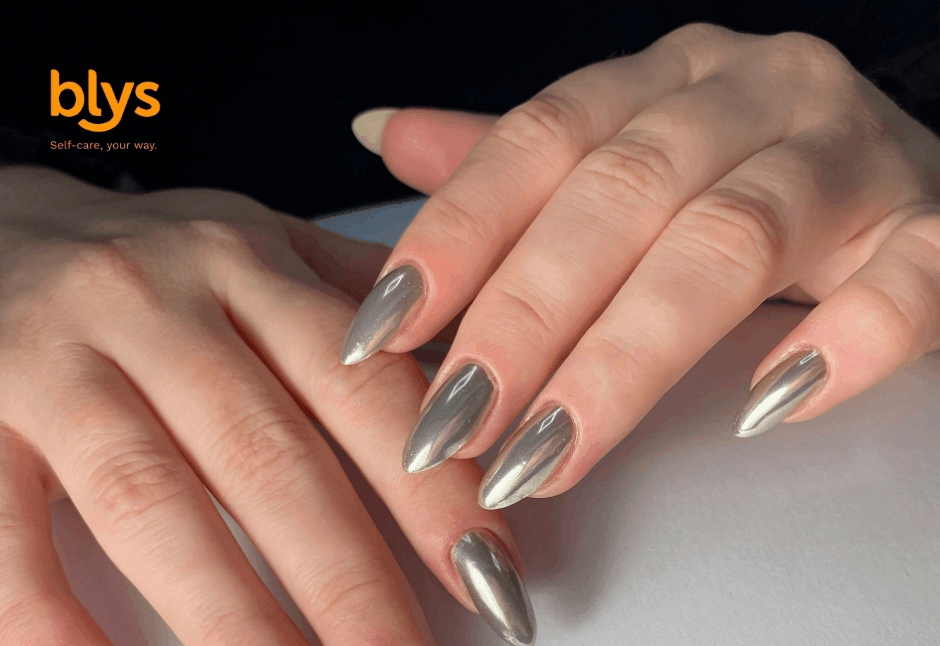 Close-up of hands with long, almond-shaped metallic silver chrome nails resting on a pale surface, showcasing a sleek, polished manicure.