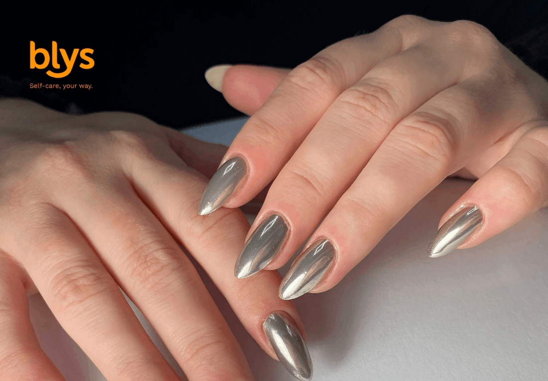 Close-up of hands with long, almond-shaped metallic silver chrome nails resting on a pale surface, showcasing a sleek, polished manicure.