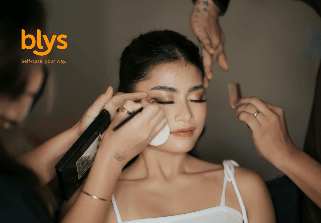 A makeup artist applies eyeliner to a serene woman while stylists adjust her hair; blys logo visible.