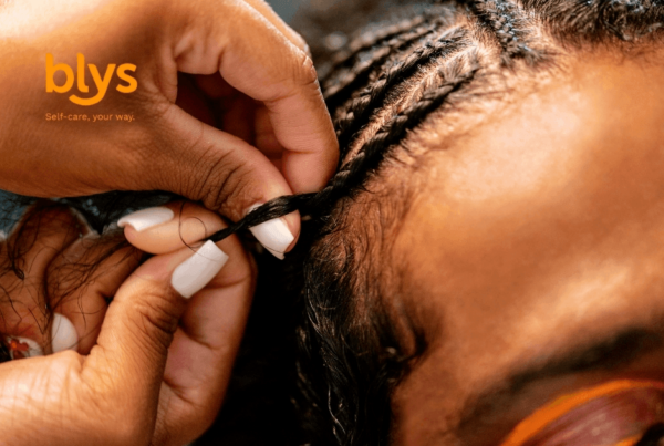 Close-up of hands braiding a Black person’s hair into neat cornrows, with white manicured nails and orange blys branding; warm, intimate beauty scene.