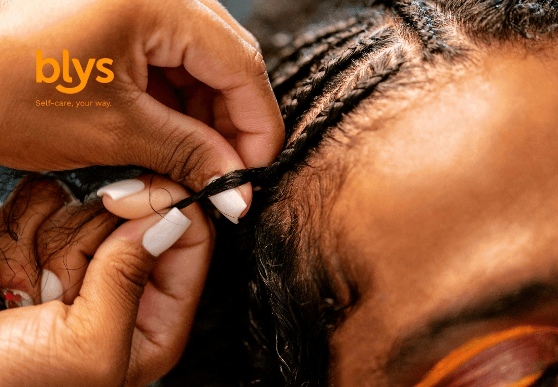 Close-up of hands braiding a Black person’s hair into neat cornrows, with white manicured nails and orange blys branding; warm, intimate beauty scene.