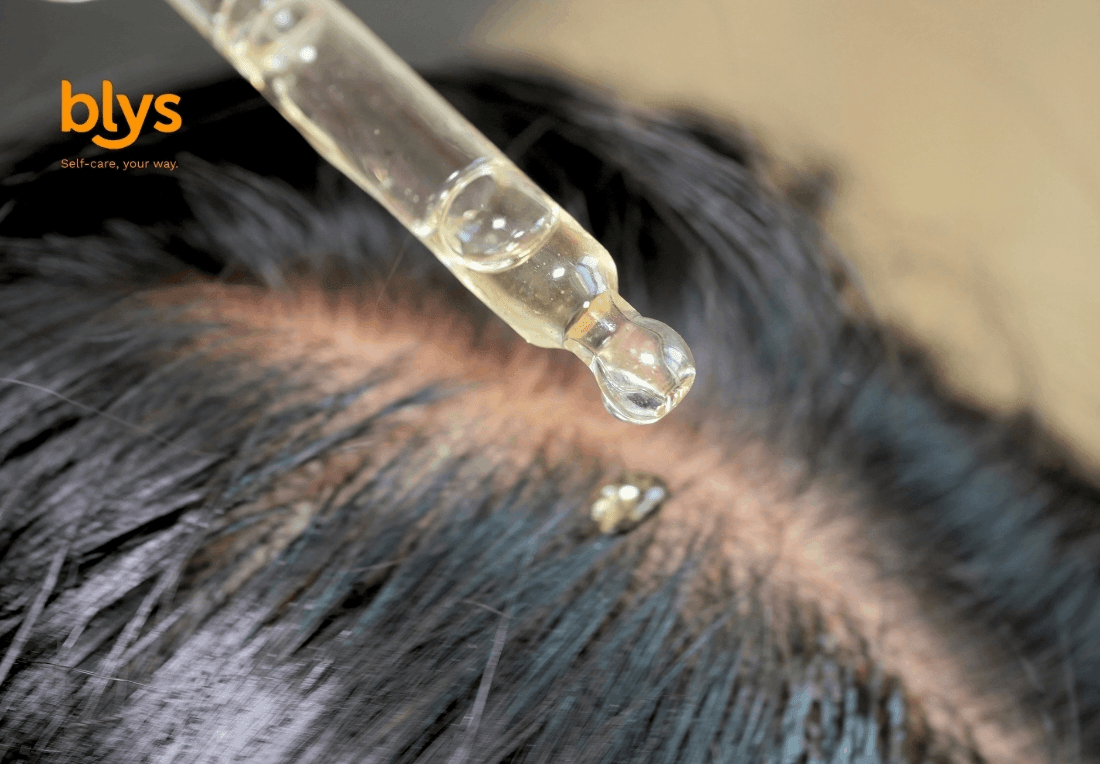 Close-up of a dropper releasing clear serum onto an exposed scalp between dark hair strands, suggesting a focused, soothing scalp treatment.