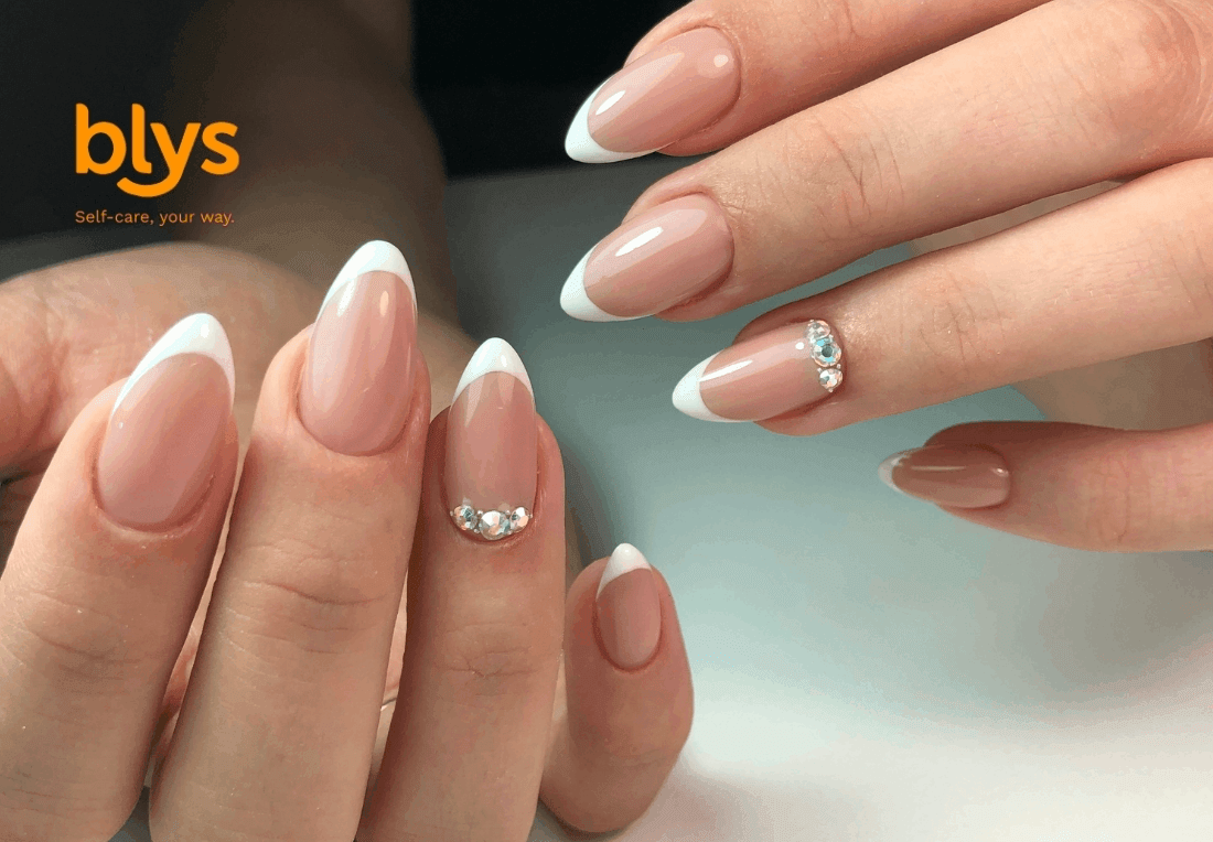 Close-up of gel manicured hands with elegant pink-and-white French-tip nails, rhinestone accents, and a blys self-care logo; polished, glamorous mood.