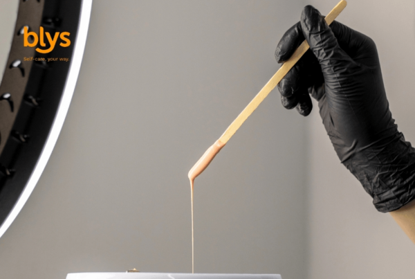Gloved hand uses a wooden stick to drizzle wax into a container, with a ring light and orange “blys” self-care logo.