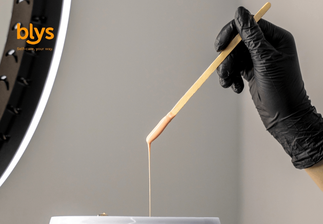 Gloved hand uses a wooden stick to drizzle wax into a container, with a ring light and orange “blys” self-care logo.