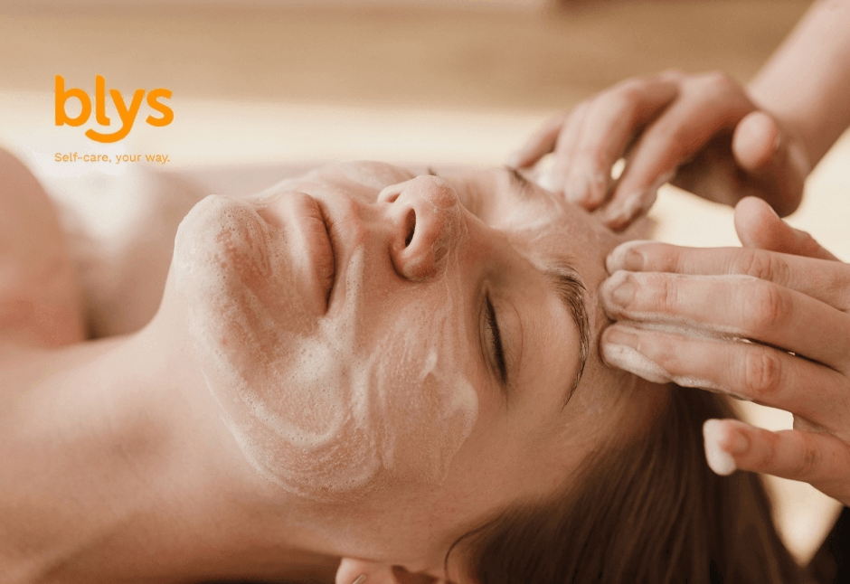 Relaxed woman receives a soothing facial massage with creamy cleanser as a skincare professional gently works across her forehead; blys logo visible.