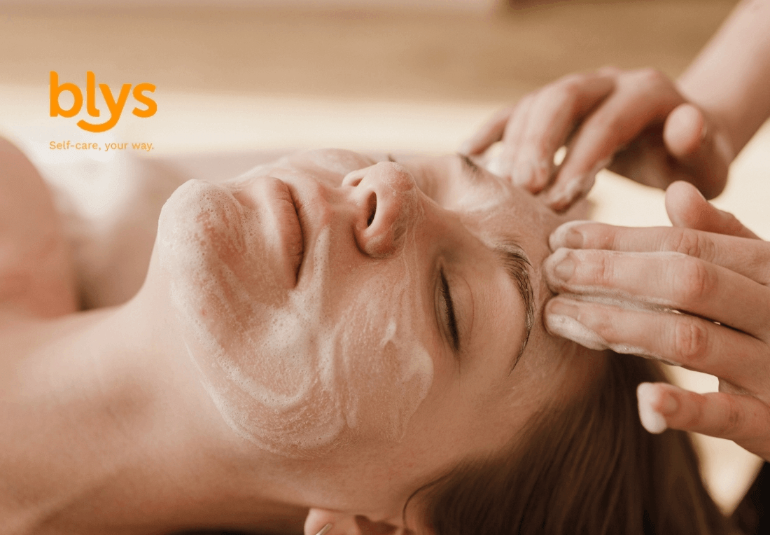 Relaxed woman receives a soothing facial massage with creamy cleanser as a skincare professional gently works across her forehead; blys logo visible.