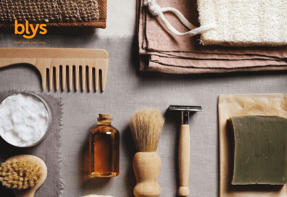 Flat lay of natural grooming essentials: wooden comb, shaving brush and razor, amber oil bottle, soap, towels, and exfoliating brush.