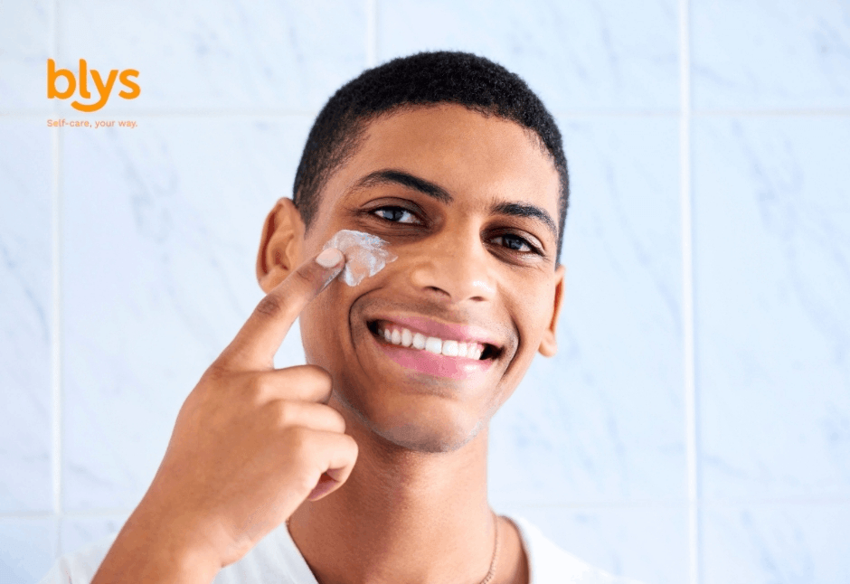 Smiling young man applies facial cream to his cheek in a bright tiled bathroom, conveying a fresh, confident skincare routine.