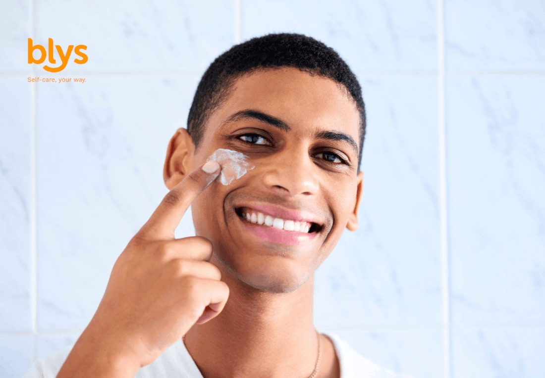 Smiling young man applies facial cream to his cheek in a bright tiled bathroom, conveying a fresh, confident skincare routine.