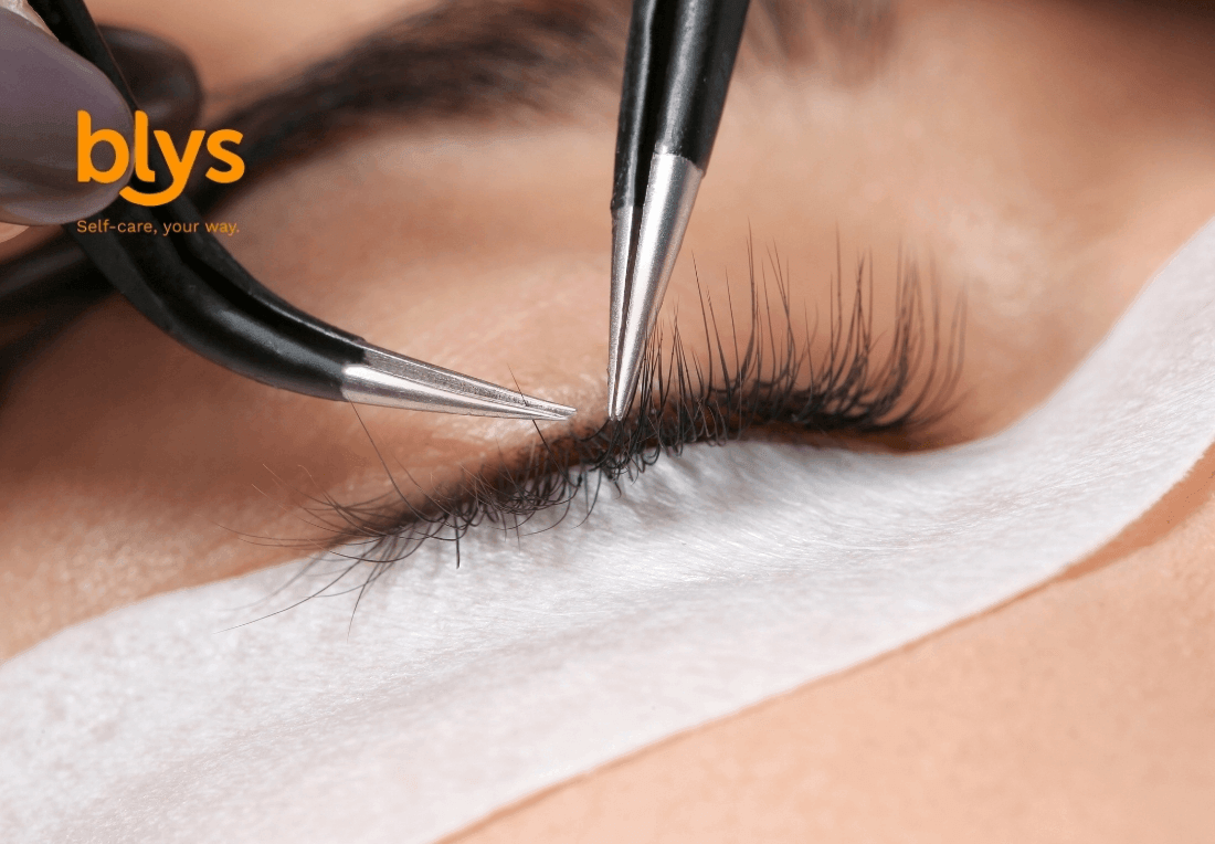 Close-up of a closed eye during eyelash extension treatment, with tweezers isolating lashes above a white under-eye pad; precise, calm beauty care.