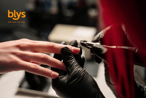 Close-up of a nail technician in black gloves applying Russian manicure to a client’s fingernail, with a red salon cape and “blys” logo visible; professional, calm mood.