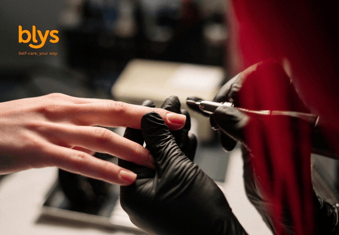 Close-up of a nail technician in black gloves doing Russian manicure to a client’s fingernail, with a red salon cape and “blys” logo visible; professional, calm mood.