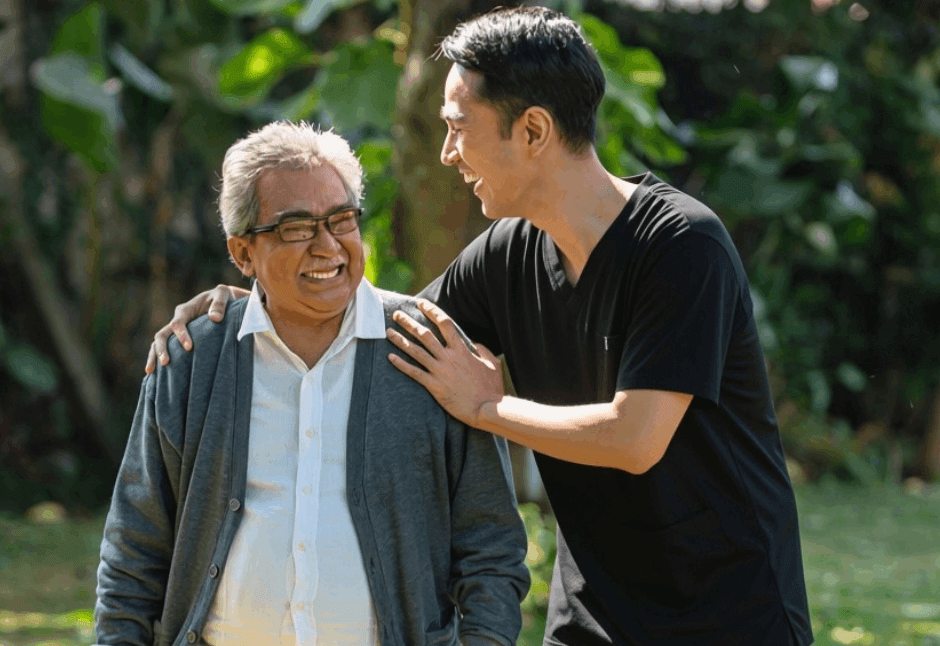 A younger man in a black shirt rests a hand on an older man's shoulder as they smile together in a leafy park, conveying warmth and affection.