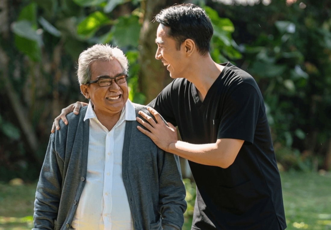 A younger man in a black shirt rests a hand on an older man's shoulder as they smile together in a leafy park, conveying warmth and affection.