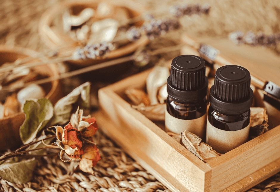 A warm, rustic arrangement of two dark essential-oil bottles in a wooden tray, surrounded by dried flowers, herbs, and natural aromatherapy massage scents.