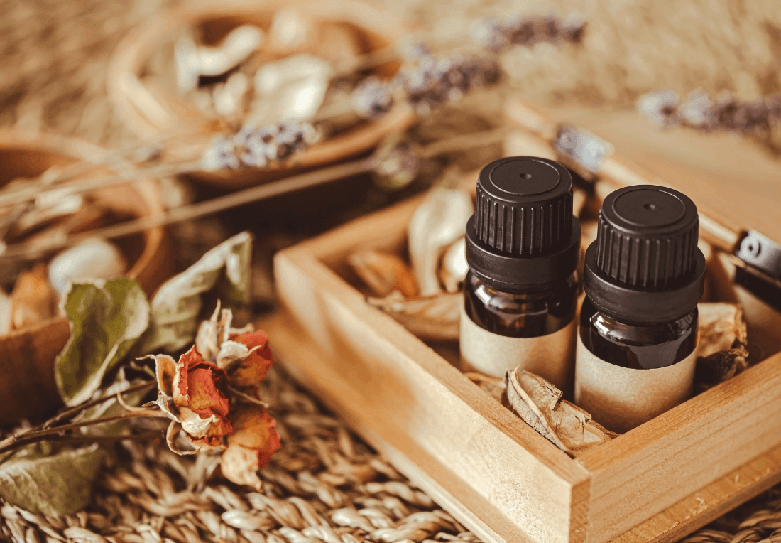 A warm, rustic arrangement of two dark essential-oil bottles in a wooden tray, surrounded by dried flowers, herbs, and natural aromatherapy massage scents.