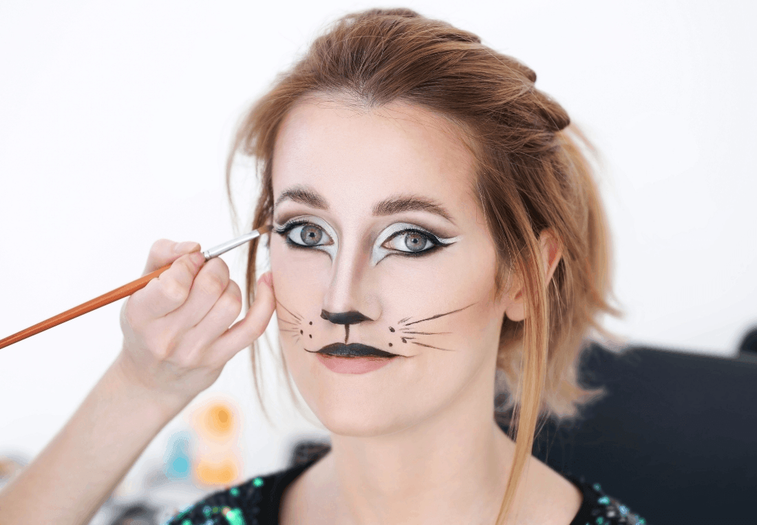 A makeup artist paints whiskers beside a woman’s nose; cat-style eyeliner, a black nose, and drawn whiskers complete her playful cat look.