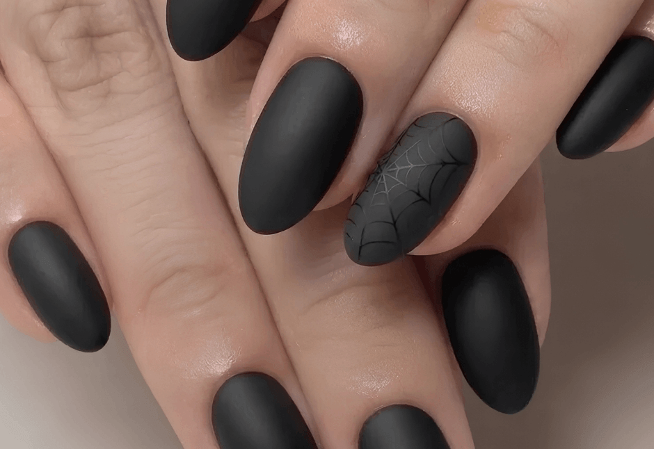 Close-up of manicured hands with matte black nails and a spiderweb accent nail, creating a sleek, moody Halloween look.