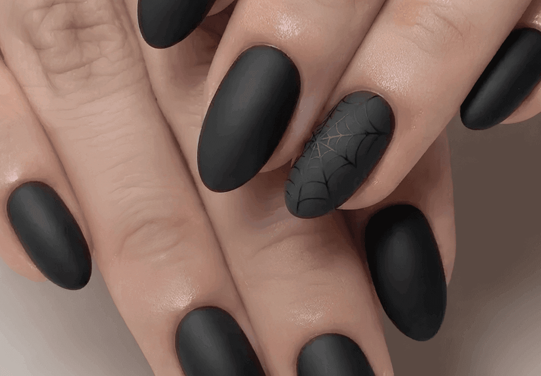 Close-up of manicured hands with matte black nails and a spiderweb accent nail, creating a sleek, moody Halloween look.