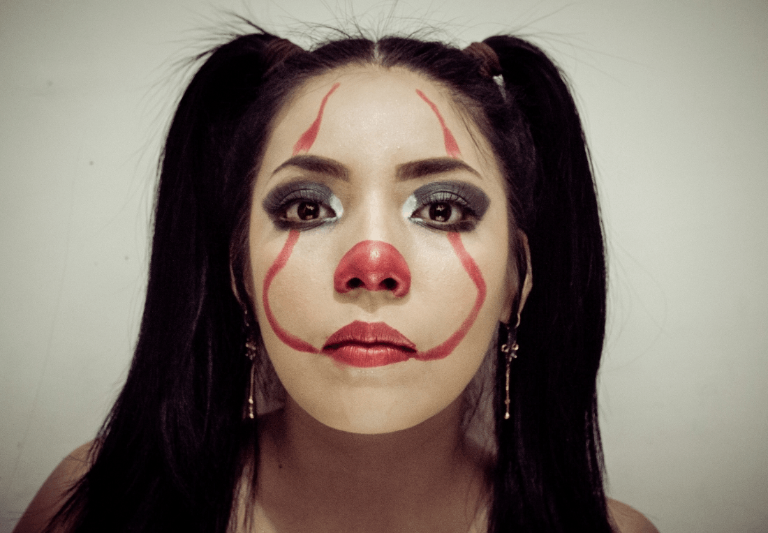 Close-up portrait of a woman with black pigtails and clown makeup; her neutral gaze feels somber and unsettling.