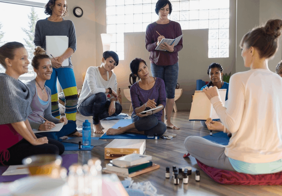 A diverse group of women coworkers sit in a sunlit studio, listening to a yoga practitioner with a clipboard; art supplies and notebooks suggest a warm, corporate retreat.