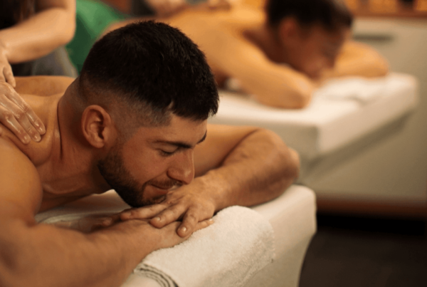 A relaxed couple receives a back massage on a white towel-covered massage table while a pair of therapists work nearby; warm lighting creates a calm, soothing mood.