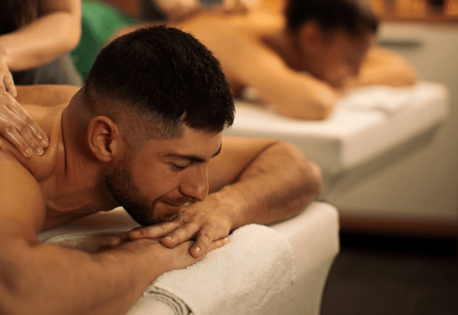 A relaxed couple receives a back massage on a white towel-covered massage table while a pair of therapists work nearby; warm lighting creates a calm, soothing mood.
