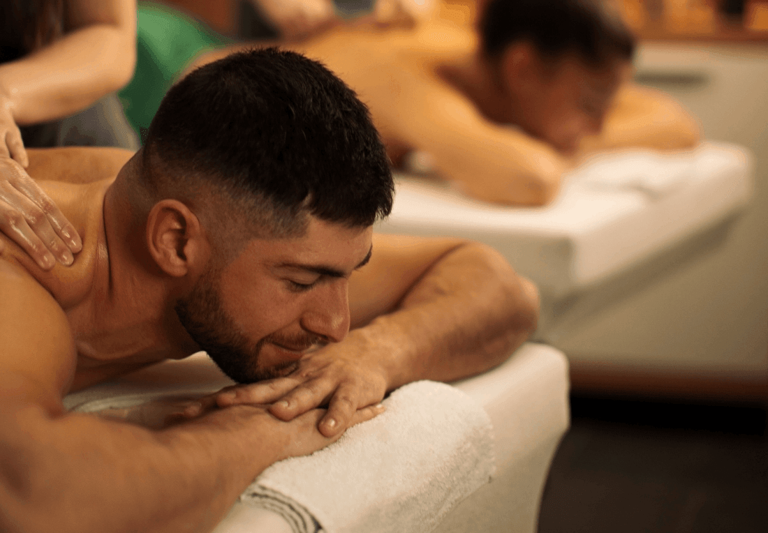 A relaxed couple receives a back massage on a white towel-covered massage table while a pair of therapists work nearby; warm lighting creates a calm, soothing mood.