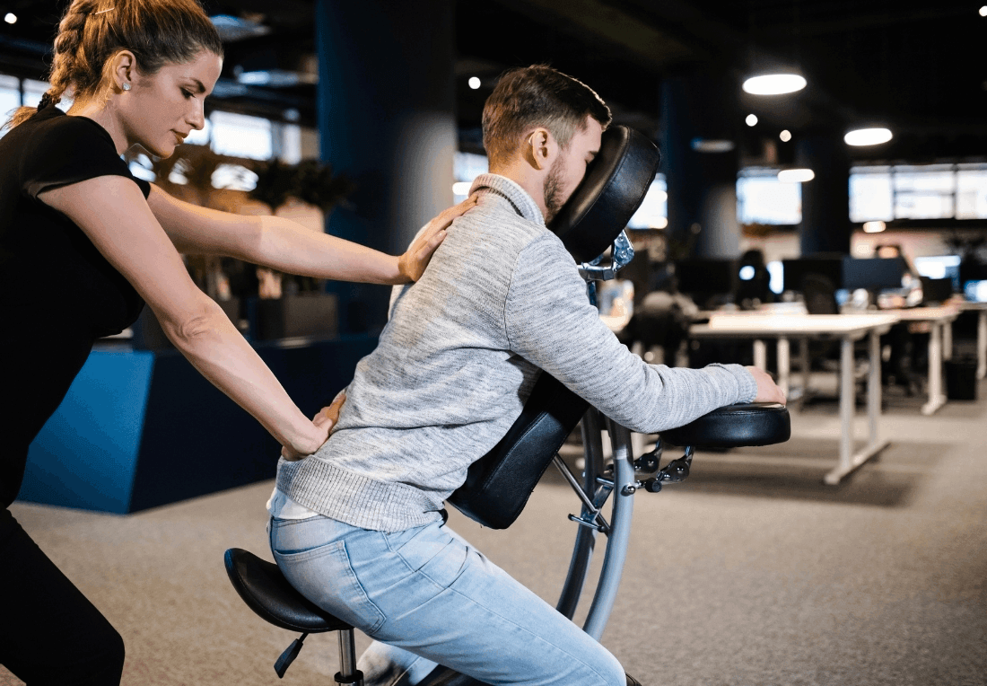 A massage therapist presses a man's back on a massage chair in a corporate setting, helping him relax during a focused, supportive chair massage session.