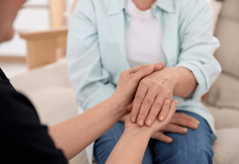 A manicure specialist gently holds an older woman’s hands in a bright room, offering comfort and support in a calm, compassionate moment.