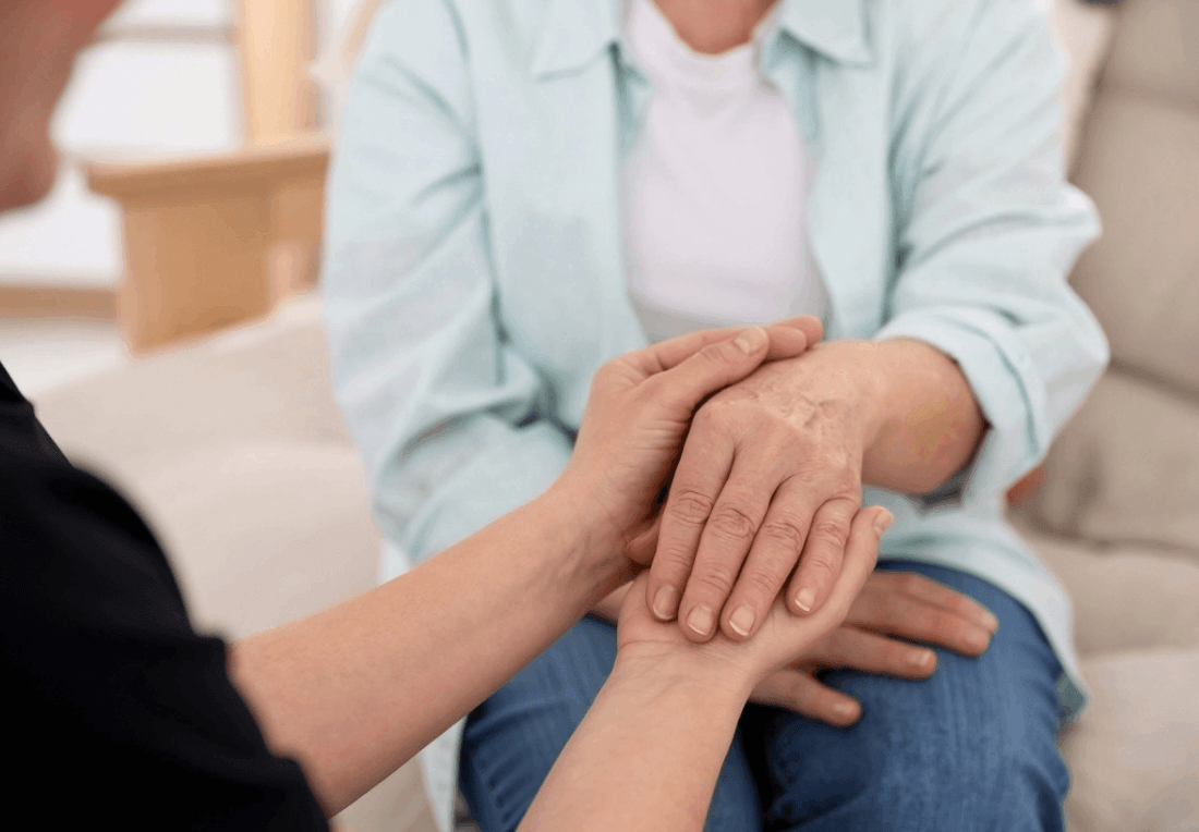 A manicure specialist gently holds an older woman’s hands in a bright room, offering comfort and support in a calm, compassionate moment.
