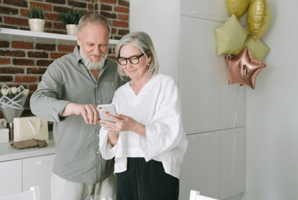 Grandparents smile as they look at a smartphone in a bright kitchen, with gold and rose-gold balloons adding a warm, celebratory feel.