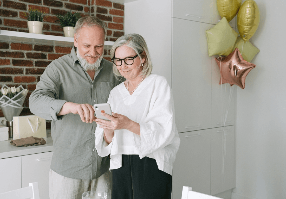 Grandparents smile as they look at a smartphone in a bright kitchen, with gold and rose-gold balloons adding a warm, celebratory feel.